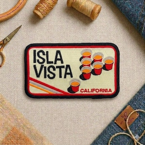 May include: Embroidered patch with the words "ISLA VISTA CALIFORNIA" in black letters on a cream background. The patch features red cups with orange liquid. The patch has a black border and red accents.