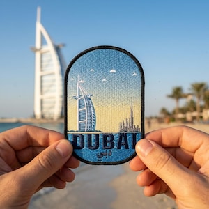 May include: An oval-shaped embroidered patch featuring a blue and yellow gradient sky background. The design depicts the Burj Al Arab and Burj Khalifa skyscrapers in white and gray thread. The word DUBAI is stitched in bold blue letters below.