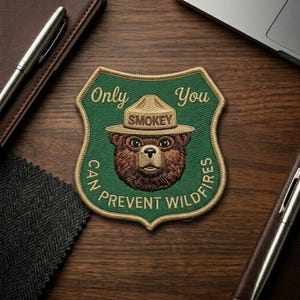 May include: Embroidered patch featuring Smokey Bear in a ranger hat, with the text "Only You Can Prevent Wildfires." The patch is shield-shaped with a green background and gold stitching. The bear's face is brown.