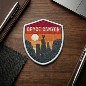 May include: Embroidered patch featuring a Bryce Canyon landscape design. The patch has a shield shape with a white border. The design includes a sunset with a sun, rock formations, and the text "BRYCE CANYON" in white.