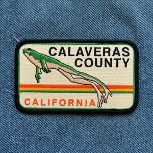 May include: Embroidered patch featuring a green frog in a stretched pose, with the words "CALAVERAS COUNTY CALIFORNIA" in black and orange text. The patch has a black border and a cream background, with orange, yellow, and green stripes.