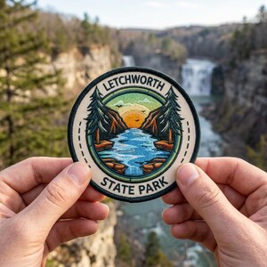 May include: Round patch with the text "LETCHWORTH STATE PARK" and a scenic illustration of a river, trees, and a sunset. The patch is held up in front of a blurred background of a waterfall and trees.