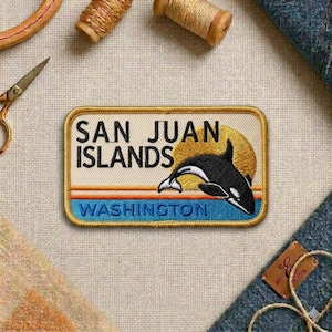 May include: Embroidered patch featuring a black and white orca leaping in front of a golden sun. The patch reads "SAN JUAN ISLANDS WASHINGTON" in a rectangular shape with a cream background and blue and orange stripes.