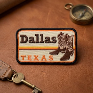 May include: Embroidered patch featuring the word "Dallas" in brown letters above the word "TEXAS" in orange. The patch includes a design of brown cowboy boots. The patch has a black border.