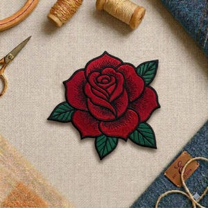 May include: A detailed, embroidered red rose patch with green leaves and black outlines. The patch is centered on a light beige fabric, with sewing tools and materials in the background, including scissors and spools of thread.