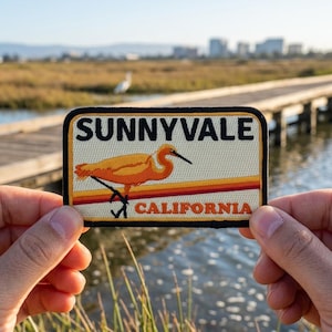 May include: A rectangular patch with the text "SUNNYVALE CALIFORNIA" and a stylized orange bird illustration. The patch has a black border and a retro color scheme of orange, red, and yellow. The patch is held up by a person.