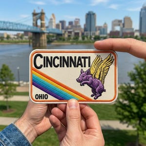 May include: A rectangular patch featuring the word "CINCINNATI" in black, with a rainbow design and a purple pig with yellow wings. The word "OHIO" is at the bottom. The patch is held up in front of a city skyline.