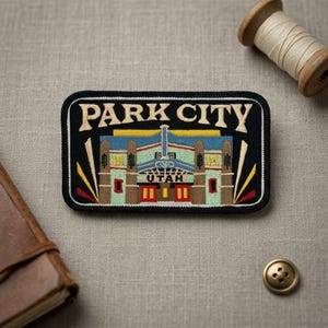 May include: Embroidered patch with the words "PARK CITY UTAH" in white lettering on a black background. The patch features a colorful illustration of a building with red, green, and blue accents. A wooden spool of thread and a button are in the background.