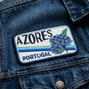 May include: A rectangular patch with the text "AZORES PORTUGAL" and a blue floral design. The patch is embroidered with a white background, blue border, and green and blue stripes. It is attached to a denim jacket.