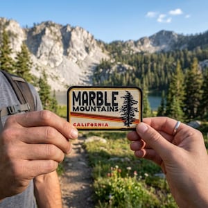 May include: A rectangular patch with the words "MARBLE MOUNTAINS CALIFORNIA" in black and red lettering. A stylized tree is on the right side. The patch is held in front of a mountain and forest landscape.