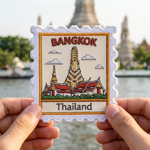 May include: A rectangular embroidered patch shaped like a postage stamp with scalloped edges. It features a cream background with the text BANGKOK in red and Thailand in black. The design depicts a golden temple with red roofs and green foliage.