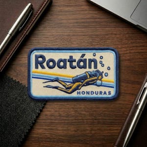 May include: Embroidered patch featuring the word "Roatán" in blue and yellow, with a diver illustration. The patch has a blue border and the word "HONDURAS" below the diver. The patch is rectangular.