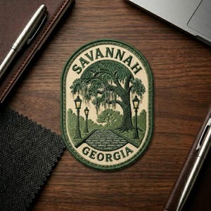 May include: Oval patch with the text "SAVANNAH GEORGIA" in green and cream. The patch features a tree, a brick path, and two street lamps. The patch is on a wooden surface with a pen and notebook.