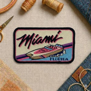 May include: Embroidered patch featuring the word "Miami" in black script above a pink and brown speedboat. The patch has a black border and includes the word "FLORIDA" below the boat, set against a cream, blue, and purple background.