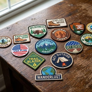 May include: A collection of embroidered patches with outdoor and travel themes. Designs include a hiking boot, a bison, the USA flag, mountains, a tent, and the words "Explore More," "Adventure Awaits," and "Wanderlust." Patches are in various shapes and colors.