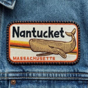 May include: Embroidered patch featuring a whale illustration with the word "Nantucket" above and "Massachusetts" below. The patch has a brown border and is set against a blue denim background.