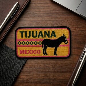 May include: Embroidered patch featuring the word "TIJUANA" above "MEXICO" with a black donkey silhouette. The patch has a yellow background with pink and green accents. A travel souvenir.