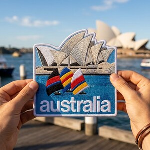 May include: A rectangular embroidered patch featuring the Sydney Opera House in white and beige tones above a blue water scene with three colorful sailboats. The word "australia" is stitched in white lowercase letters at the bottom.