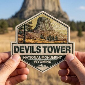 May include: Embroidered patch featuring Devils Tower National Monument in Wyoming. The patch has a detailed illustration of the tower, trees, and landscape, with the words "DEVILS TOWER" and "NATIONAL MONUMENT WYOMING" in a rectangular border.