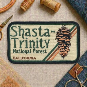 May include: Embroidered patch featuring the text "Shasta-Trinity National Forest CALIFORNIA" with a pinecone graphic. The patch has a cream background, a dark green border, and brown and green diagonal stripes.