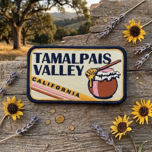 May include: A rectangular embroidered patch with a cream background and a dark blue border. It features the text TAMALPAIS VALLEY CALIFORNIA in dark blue, a honey dipper, and a jar of honey with pink and brown accents and diagonal stripes.