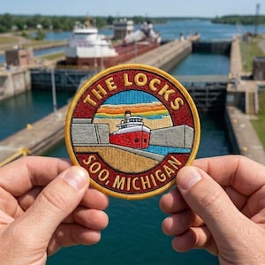 May include: A circular embroidered patch featuring a red and white ship inside a canal lock. The border is yellow with a dark red background. The text reads THE LOCKS at the top and SOO, MICHIGAN at the bottom in gold, capitalized, sans-serif letters.