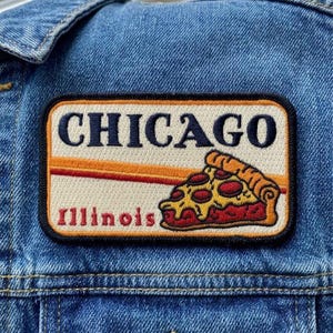 May include: Embroidered patch featuring the word "CHICAGO" above "Illinois" and a pizza slice. The patch has a cream background with orange and red stripes, and a black border. The patch is sewn onto a denim jacket.