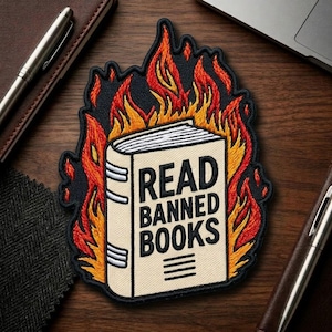 May include: Embroidered patch featuring a book engulfed in flames. The book is beige with the words "READ BANNED BOOKS" in black. The flames are red, orange, and yellow. The patch is on a dark background with pens and a laptop.