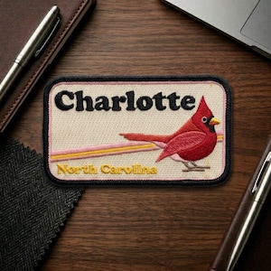 May include: Embroidered patch featuring a red cardinal bird and the text "Charlotte North Carolina". The patch has a cream background with pink and yellow stripes, and a black border. The patch is rectangular.