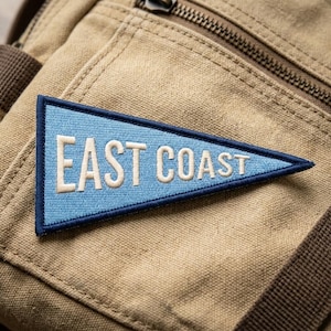 May include: A triangular pennant patch featuring a light blue background with a dark blue border. The text EAST COAST is embroidered in white capital letters across the center of the patch, which is attached to a tan canvas fabric surface.