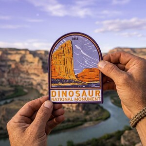 May include: A patch featuring a landscape of Dinosaur National Monument. The patch has a purple border and includes the text "DINOSAUR NATIONAL MONUMENT" and the year "1915". The image depicts a rock formation in shades of orange and yellow.