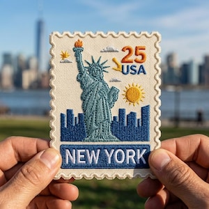 May include: Embroidered patch featuring the Statue of Liberty, a sun, and the text "25 USA" and "NEW YORK." The patch has a postage stamp design with a cream-colored background and blue and orange accents. The patch is held up in front of a blurred city skyline.