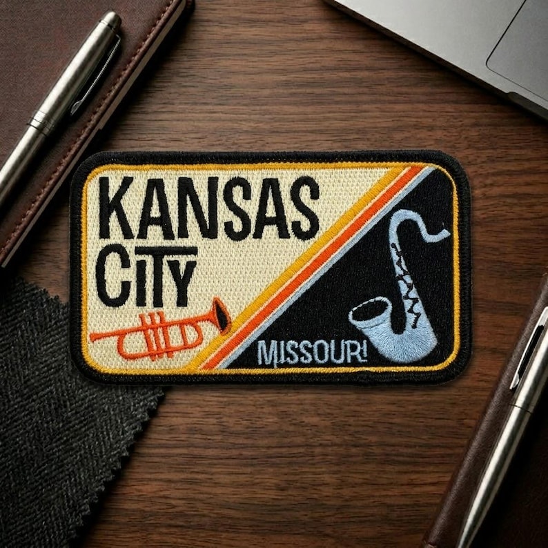 May include: Embroidered patch featuring "KANSAS CITY MISSOURI!" in black and white text. The patch includes a trumpet and a saxophone, with orange, yellow, and blue accents. The patch is rectangular with a black border.