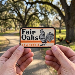 May include: A rectangular patch with the words "Fair Oaks CALIFORNIA" and a rooster illustration. The patch has a cream background with black and orange text and a black border. The patch is held between two hands.
