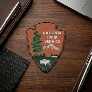 May include: Embroidered patch featuring the National Park Service logo. The patch is shaped like an arrowhead and includes a green tree, a mountain, and a bison. The text "NATIONAL PARK SERVICE" is displayed in white.