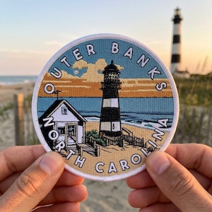 May include: A circular embroidered patch featuring a coastal scene with a black and white striped lighthouse and a small house. The text "OUTER BANKS NORTH CAROLINA" is stitched in white along the border against a blue, orange, and tan background.