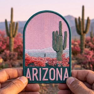 May include: An arched embroidered patch featuring a desert landscape with a saguaro cactus, pink sky, and red flora. The word ARIZONA is stitched in large pink capital letters at the bottom, all bordered by a dark teal edge on a fabric base.