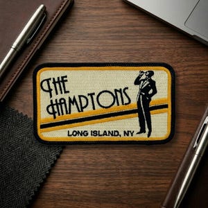 May include: Embroidered patch with the text "The Hamptons" in black and yellow, with a black and yellow striped design. A black silhouette of a person drinking is on the right. The text "Long Island, NY" is below.