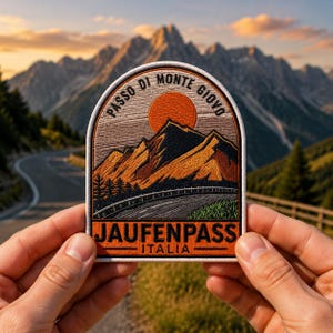 May include: Embroidered patch featuring a mountain landscape with a road and a setting sun. The patch reads "Passo di Monte Giovo" and "Jaufenpass Italia" in an arched design. The patch is held in front of a blurred mountain backdrop.