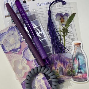 Puede incluir: Una colección de artículos de papelería morados, que incluyen bolígrafos, un marcapáginas, una goma para el pelo y pegatinas. Los artículos están dispuestos sobre una superficie blanca. El marcapáginas tiene una borla y una flor prensada. Los bolígrafos están etiquetados "erasable gel pen".