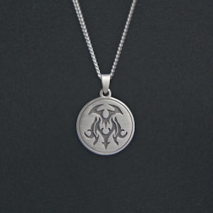 Puede incluir: Collar con colgante plateado con un colgante circular con un diseño tribal grabado. El colgante cuelga de una delicada cadena. El fondo es de color oscuro.