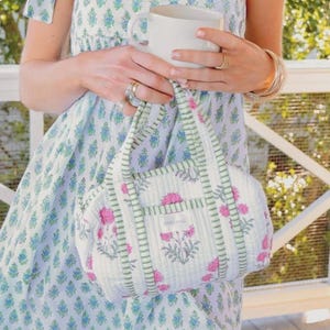 Puede incluir: Un bolso acolchado con base blanca, patrón de rayas verdes y motivos florales rosas. El bolso incluye dos asas superiores y una pequeña etiqueta rectangular en la parte frontal con el texto DARLING impreso en letras mayúsculas negras.