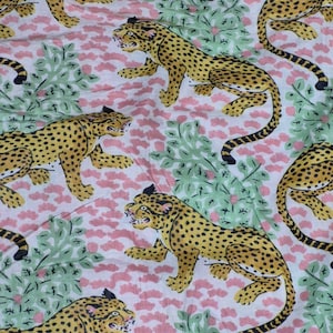 May include: A repeating pattern of yellow leopards with black spots and tails on a white background. The leopards are interspersed with pink floral and green leafy elements. The design has a vintage, jungle-inspired aesthetic.