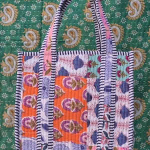 May include: A patchwork tote bag with a variety of floral patterns in orange, blue, and white. The bag has a striped border and handles, and is held against a green background with a paisley design.