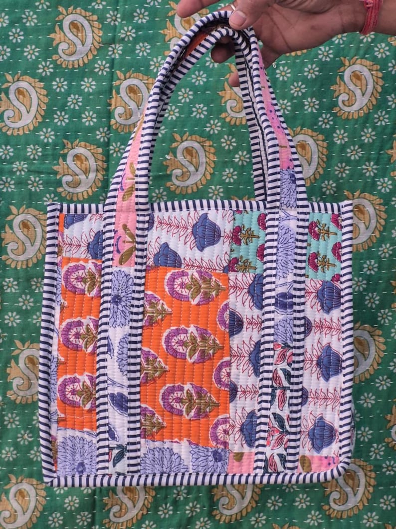 May include: A patchwork tote bag with a variety of floral patterns in orange, blue, and white. The bag has a striped border and handles, and is held against a green background with a paisley design.