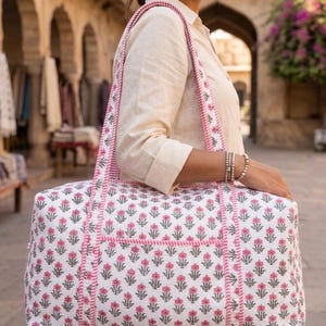 May include: A rectangular quilted duffel bag featuring a white base with a repeating pattern of small pink flowers and grey leaves. The bag includes two long shoulder straps and trim details in a pink and white striped fabric pattern.