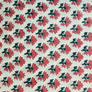 May include: White fabric with a repeating pattern of pink flowers and green leaves. The floral design is evenly spaced across the fabric, creating a visually appealing and symmetrical arrangement. The fabric appears to be cotton or a similar material.