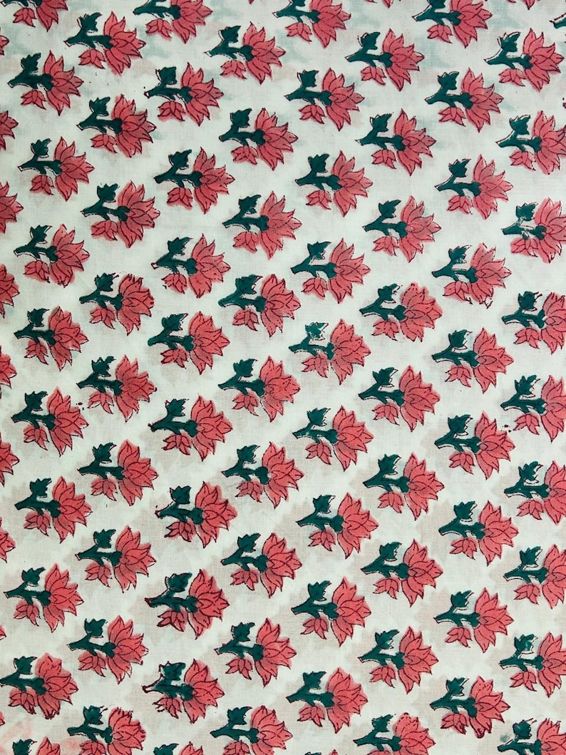 May include: White fabric with a repeating pattern of pink flowers and green leaves. The floral design is evenly spaced across the fabric, creating a visually appealing and symmetrical arrangement. The fabric appears to be cotton or a similar material.