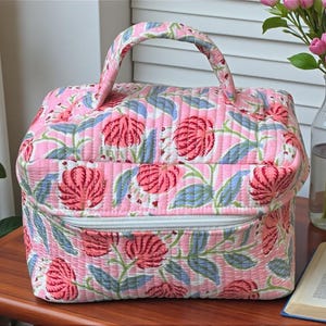 May include: A pink quilted bag with a floral pattern featuring red flowers and blue leaves. The bag has a white zipper, a matching handle, and is sitting on a wooden table. The bag is approximately 10 inches wide and 8 inches tall.