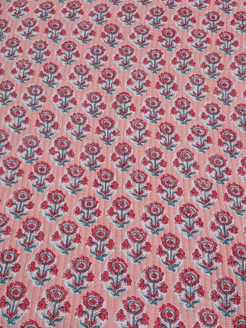 May include: A quilted fabric with a repeating floral pattern. The flowers are red with white and teal accents, set against a light coral background. The fabric has a stitched, textured appearance.